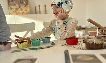 Ein Kind mit Kochmütze auf sitzt am Tisch mit bunten Backformen