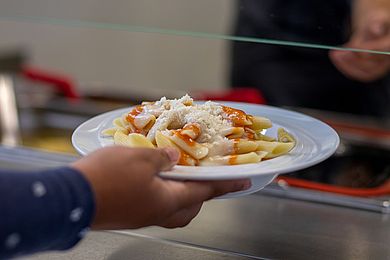 Ein Teller Nudeln mit Tomatensoße und Parmesan wird vom M9-Mitarbeitenden an den Gast ausgehändigt