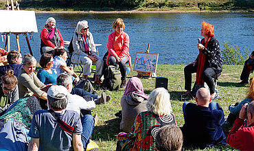 Mehrere Menschen sitzen an einem Flussufer und hören einer erzählenden Person zu