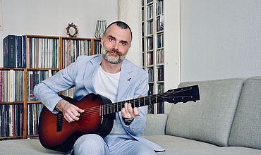 Der Musiker Ronny Valdorf sitzt auf einem Sofa und spielt eine Akusitik-Gitarre. Er trägt einen hellblauen Anzug.