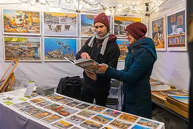 Stand mit bunten Bildern, verschiedene Motive. Zwei Personen unterhalten sich über ein Bild