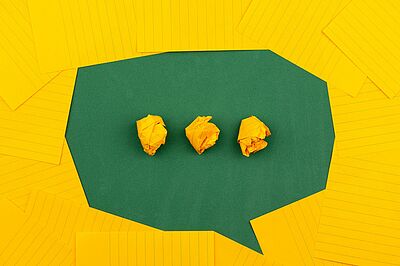 Aus Papier geformte grüne Sprechblase auf gelbem Hintergrund. In der MItte der Sprechblase drei zusammengeknüllte gelbe Papiere als "Punkte" nebeneinander drapiert.