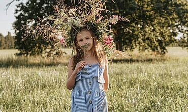 Mädchen in blau-weiß gestreiften Kleid steht auf einer grünen Wiese vor einem Baum und trägt einen großen Blumenkranz auf dem Kopf
