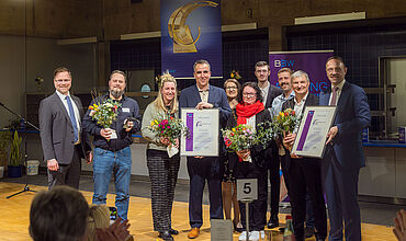 Gruppenfoto nach der Preisvergabe der Brückenschlag-Preise. Alle Preisträger*innen und die Geschäftsführung der BBW-Leipzig-Gruppe mit Blumensträußen, den Preisen und den Urkunden auf der Bühne.