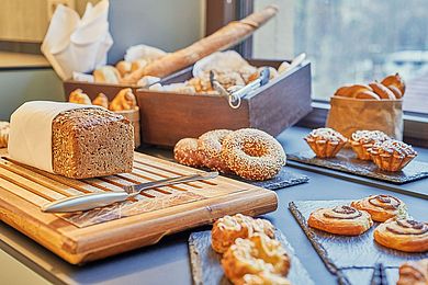 Ein in ein Baumwolltuch geschlagenes Vollkornbrot mit Sonnenblumenkernen, neben dem ein großes Messer liegt, wartet auf den Anschnitt beim Frühstücksbuffet. Dahinter sind noch weitere Brotsorten, wie zum Beispiel ein Baguette, Croissants und Bagels zu sehen. Süße Blätterteigteilchen und Leipziger Lerchen werden ebenfalls angeboten.