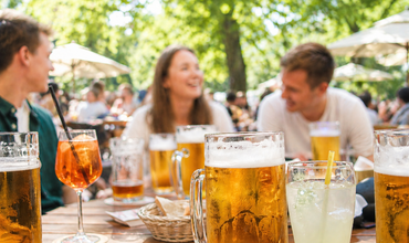verschiedene Getränke im Vordergrund. Im Hintergrund sitzen 3 Personen und sind unscharf zu erkennen. Die Stimmung ist fröhlich, das Licht sommerlich.